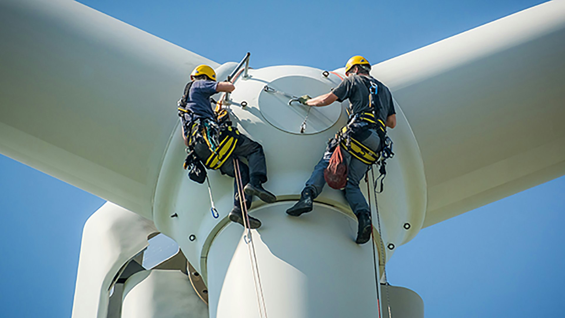 Two people in harnesses work on the hub of a wind turbine