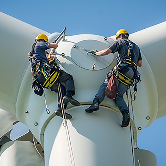 Two people in harnesses work on the hub of a wind turbine